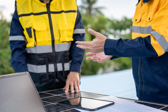 Two solar energy technicians in safety uniforms discuss photovoltaic panels with a laptop and tablet during an outdoor site inspection.