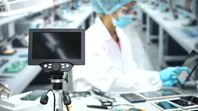 Digital microscope monitor in front of a masked technician inspecting electronic circuit boards on a clean factory workbench.