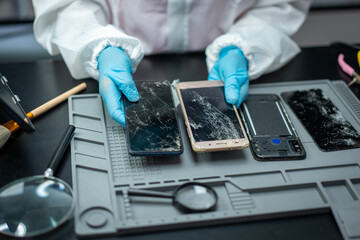 Technician in protective gloves compares cracked smart phone screens on a repair mat with tools, showing mobile device damage and electronics service. © NanSan