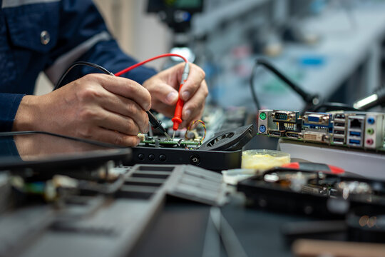 Close-up of an electronics technician testing a computer circuit board with multimeter probes at a repair workbench.