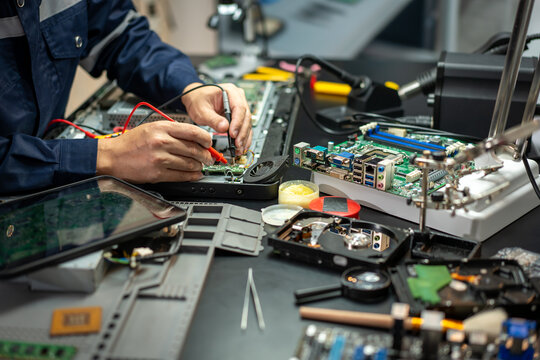 Electronics technician repairs an open laptop motherboard with test probes at a busy workbench filled with computer parts, tools, and hard drives.