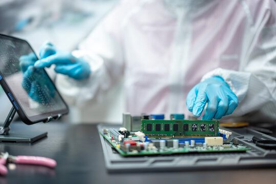 Technician in protective clothing and blue gloves repairing a computer circuit board with memory module at an electronics workbench.