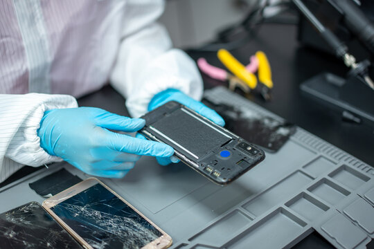 Technician wearing blue gloves repairs an opened smart phone with a cracked screen on a workbench, using tools and spare parts.