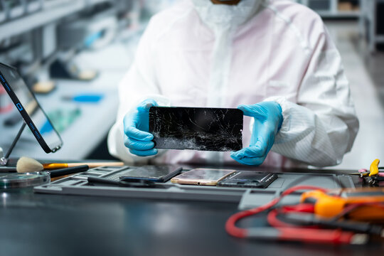 Technician in protective clothing and blue gloves holds a cracked smart phone screen above a workbench with repair tools and damaged phones.