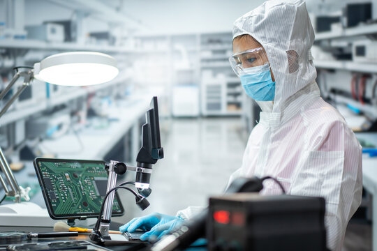 Electronics technician in clean room clothing inspects a circuit board with a microscope in a modern laboratory.