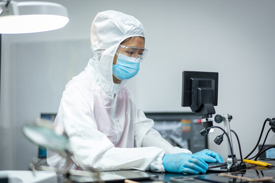 Technician in clean room suit, mask and safety glasses repairing electronic equipment at a laboratory workbench.