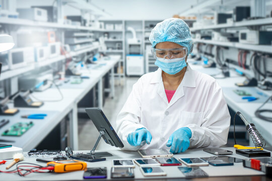 Technician in protective clothing repairs smart phones on a workbench in a modern electronics laboratory.