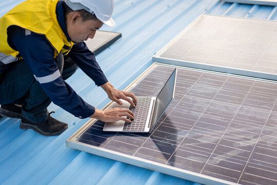 Solar technician in safety gear uses a laptop beside rooftop solar panels, checking a renewable energy system on a sunny day.