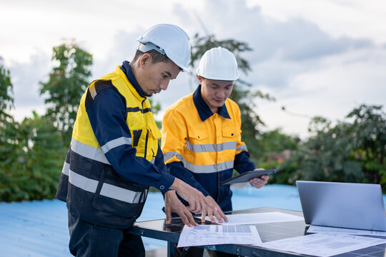 Two construction engineers in safety helmets and reflective work wear review blueprints beside a laptop at an outdoor work site.