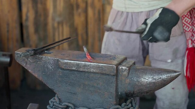 Adult blacksmith holds heated metal against anvil with protective gloves and concentrated expression. Workshop trainee practices careful shaping beside rustic timber walls in forge setting.
