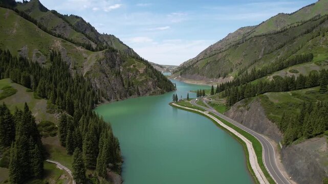 Aerial drone view of turquoise alpine lake nestled in lush green mountain valley with winding road and conifer forest, Tianshan Grand Canyon scenic area
