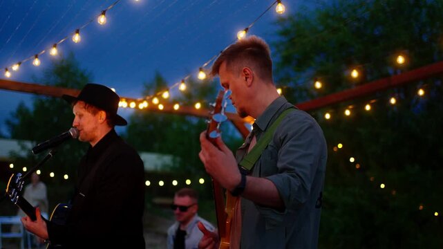 Live outdoor band performance under string lights at twilight with guitarist and singer playing music on stage during an intimate evening gathering