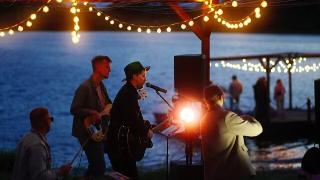 Outdoor lakeside evening concert with live band performing under glowing string lights as audience gathers by the water and boats rest along the illuminated dock