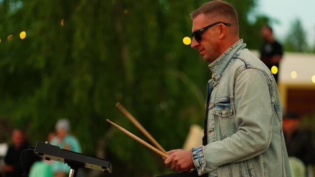 Outdoor musician playing electronic drums in denim jacket and sunglasses at a relaxed evening gathering with blurred people and warm festival lights behind