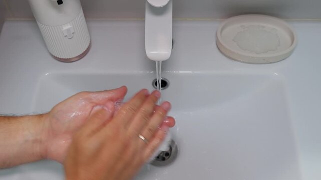 Adult hands wash under faucet in clean hygienic routine, rubbing soap into skin before rinsing at bathroom sink. Careful hand washing continues with steady motion and white ceramic basin.