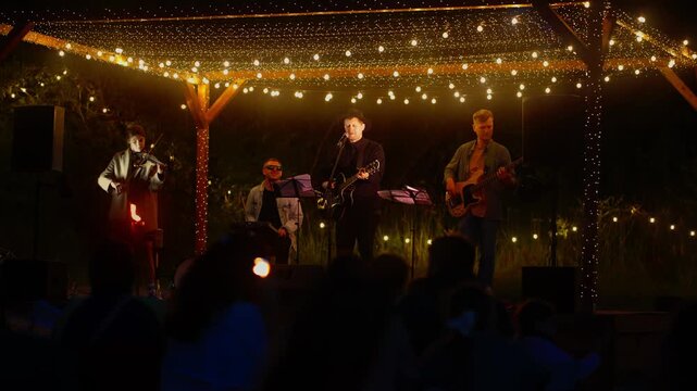 Intimate outdoor evening concert with live band performing under warm string lights on rustic wooden stage surrounded by nighttime crowd and soft glowing ambiance