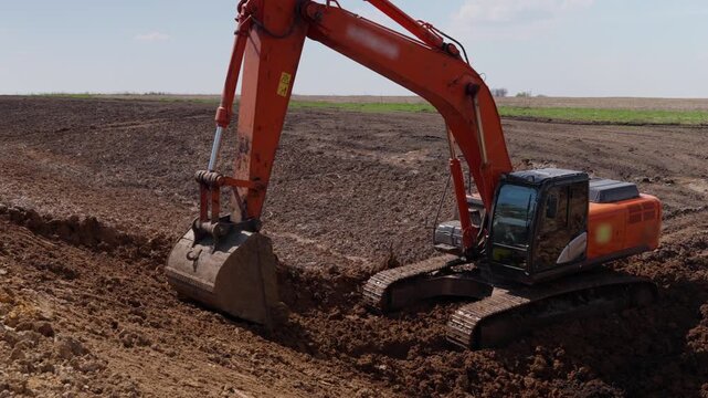 Excavator cutting soil in wide field with steady control and practical earthmoving work. Orange machine lowers bucket, lifts earth, forms rough trench, and shifts soil through repeated digging.