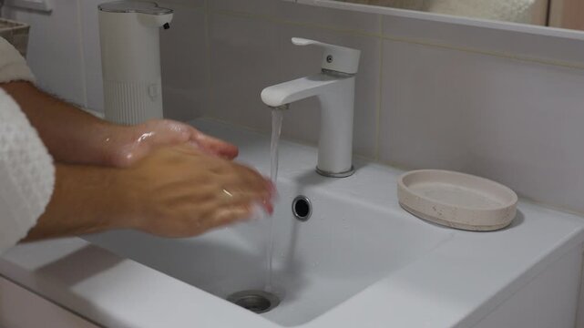 Adult man washes hands beneath running bathroom faucet with concentrated expression. Male handwashing continues as soap lather covers palms and fingers during hygiene routine at bathroom sink.