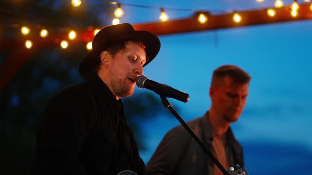Outdoor evening acoustic concert with male singer in black hat performing live under warm string lights against twilight sky, playing guitar for intimate audience