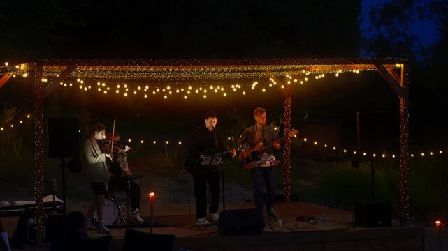 Intimate outdoor night concert with acoustic musicians performing under warm string lights on a small rustic wooden stage surrounded by nature