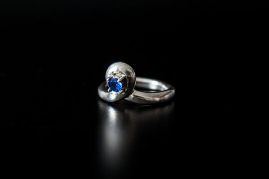 A silver ring with a blue gemstone sits on a black surface. The ring has a smooth, polished finish and a simple design, emphasizing the gemstone's color.