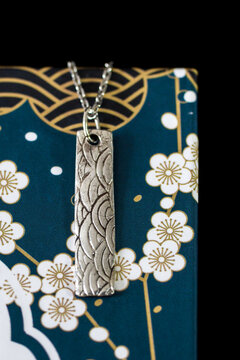 A silver pendant with intricate wave patterns hangs on a chain against a blue background adorned with white flowers and gold accents.