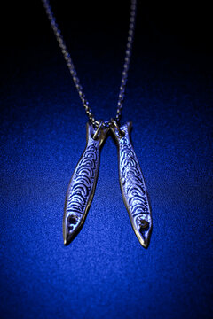 A silver necklace featuring two fish-shaped pendants with intricate designs. The background is a deep blue, enhancing the jewelry's shine.