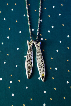 Silver fish-shaped pendant necklace on a teal background with gold polka dots. The necklace features two fish charms with intricate designs.