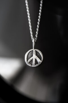 A silver peace symbol pendant hangs from a delicate chain. The background is softly blurred, emphasizing the pendant's design and shine.