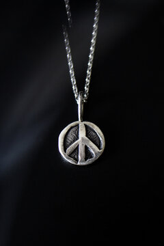 A silver peace symbol pendant hangs from a delicate chain against a dark background. The pendant features a classic design representing harmony and unity.
