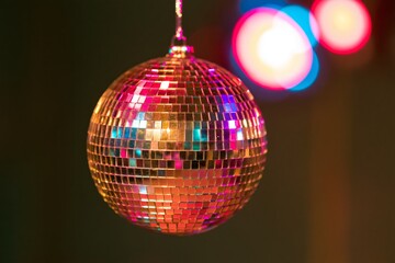 Obraz premium Closeup of a faceted mirrored disco ball suspended against dark background with colorful bokeh lights reflecting magenta cyan and golden highlights across its surface creating vibrant festive