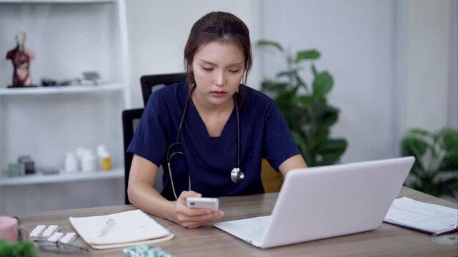 Young Asian doctor checking smartphone while working on laptop at clinic office desk