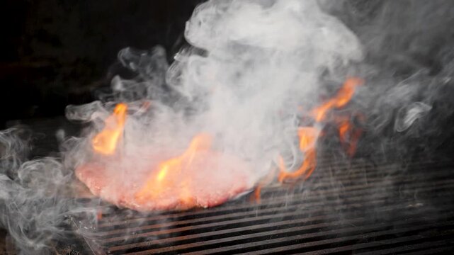 Juicy beef patty grilling on metal grate with intense orange flames and thick smoke rising. Dramatic dark background, professional restaurant kitchen cooking process. Slow motion 4k video