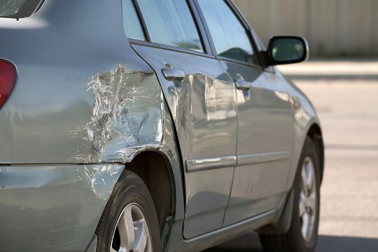 Close-up side view of metallic gray passenger car showing deep dent and scraped paint on rear door panel under neutral daylight with intact front and rear sections visible in plain outdoor parking