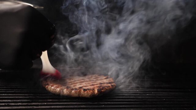 Chef hand in black glove grilling beef patty on metal grate with intense orange flames and thick smoke. Dramatic dark background, professional restaurant cooking process Slow motion 4k video