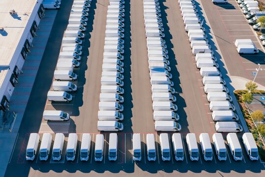 Aerial drone view above rows of identical white delivery vans parked in precise parallel lines at a large commercial depot with loading buildings and marked parking zones under bright morning