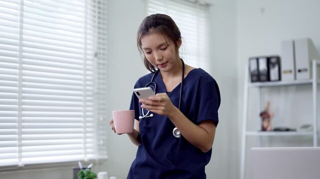 Young Asian doctor holding coffee mug checking smartphone during break in bright medical office