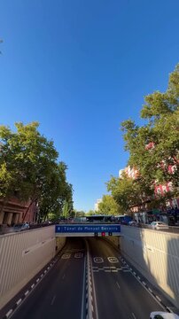 Urban traffic flowing through the tunnel under Manuel Becerra square in Madrid, Spain. A typical city scene with cars, buildings, and leafy trees lining the avenue under a clear blue summer sky