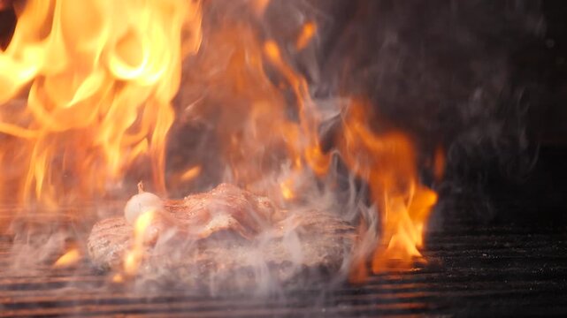 Juicy beef patty grilling on metal grate with intense orange flames and thick smoke rising. Dramatic dark background, professional restaurant kitchen cooking process. Slow motion 4k video