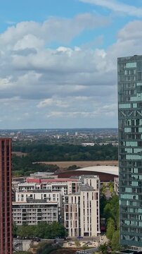 Drone view of Manhattan loft gardens in Stratford, London, panning right to reveal the distinctive notch cutout in a modern high-rise, with skyline and clouds Vertical format.