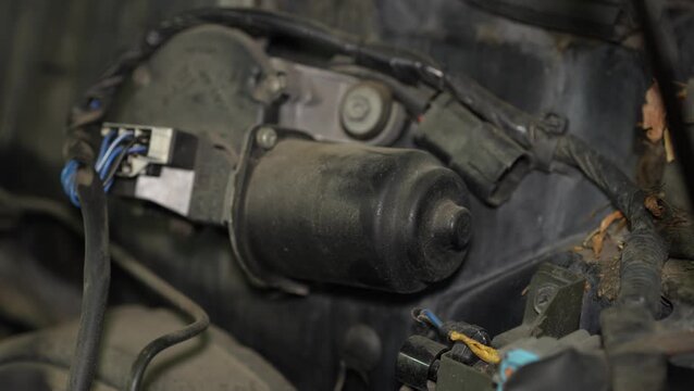 A dusty black electric windshield wiper motor assembly with electrical connectors and wiring harness is mounted in a car engine bay.