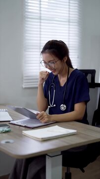 Young Asian female doctor reviewing patient records on tablet at desk in bright clinic office