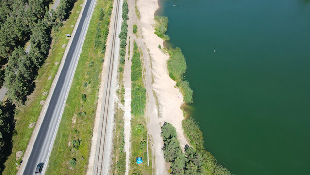 Drone view captures a vibrant green lake with a sandy beach, alongside parallel railroad tracks and a highway, demonstrating the coexistence of nature and infrastructure