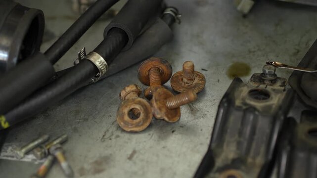 Several rusty metal bolts and washers lie on a greasy workshop table during automotive engine repair and disassembly.