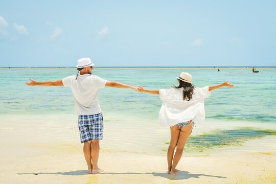 Caucasian couple man woman stand together arms outstretched enjoy white sand beach in South Zanzibar, Tanzania. Romantic summer holiday by turquoise ocean with clear blue sky. Honeymoon paradise vibes