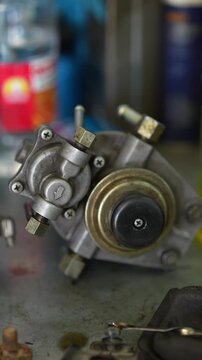 A removed high-pressure fuel pump with a mechanical priming button sits on a workbench during car engine repair work.