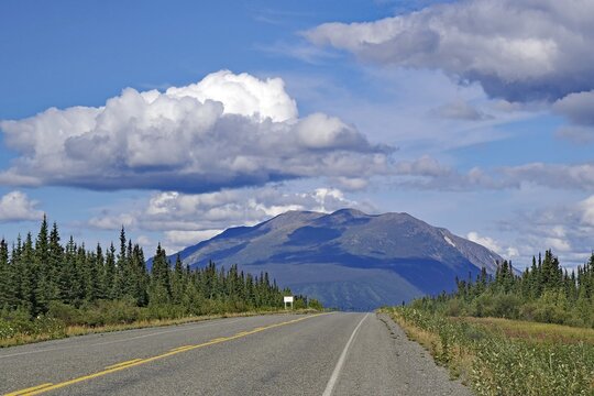 An empty road leads through a picturesque landscape with mountains and clouds in the blue sky, Alaska Highway, Yukon Territory, Canada