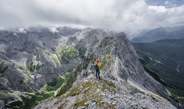 Mountaineer at the summit of the Waxenstein, view over rocky and narrow ridge of the Waxenstein ridge to H&ouml;llental with Jubil&auml;umsgrat, cloudy rocky mountain landscape, Wetterstein Mountains, Garmisch-Partenkirchen, Werdenfelser Land, Upper Bavaria, Bavaria, Germany