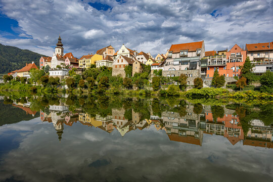 Scenic view of the historic town skyline of Frohnleiten with its parish church tower and colorful riverside buildings reflected in the Mur River under a cloudy sky in Styria, Austria