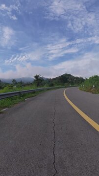 Scenic winding mountain road with a yellow center line, guardrail and lush green hills beneath a blue sky with soft clouds.